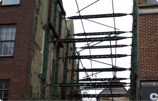 A partially demolished building with exposed steel beams and green metal supports bracing the remaining walls. The brickwork and window frames of adjacent buildings are visible on either side, with the sky and some rooftops in the background.